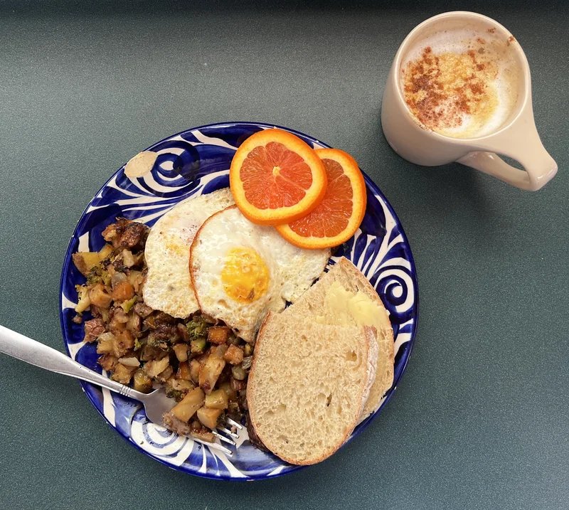 A café plate of fried eggs, hash and toasted bagel with orange slices, beside a latte.